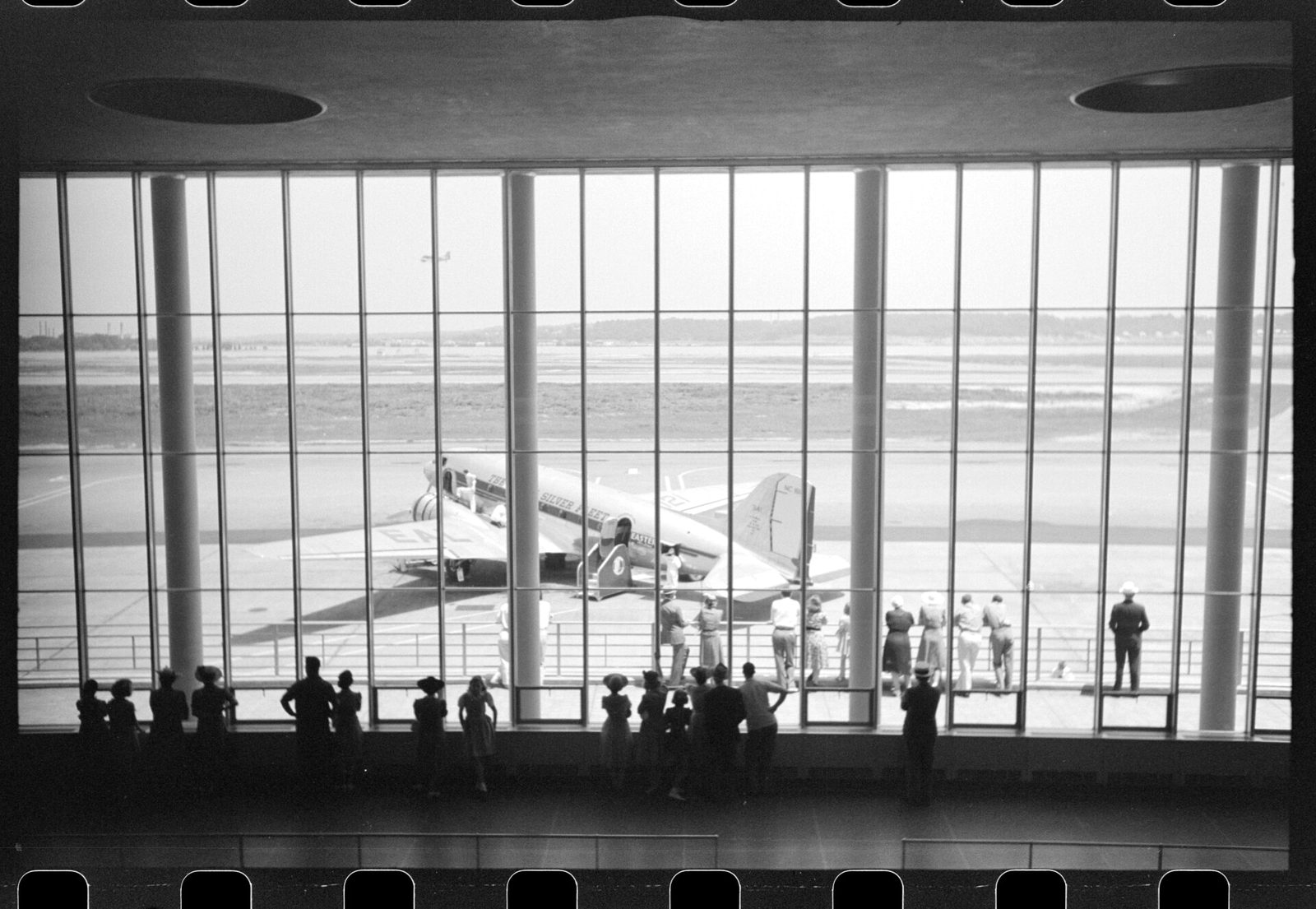 Passengers at the windows of the terminal waiting room at Washington National Airport in 1941, an airliner parked on the apron outside.